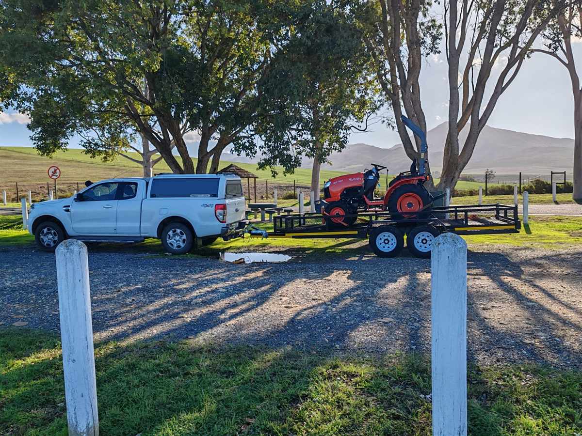 Branded white Ford Ranger bakkie with a Kubota tractor on a flatbed trailer, parked under trees at golden hour.