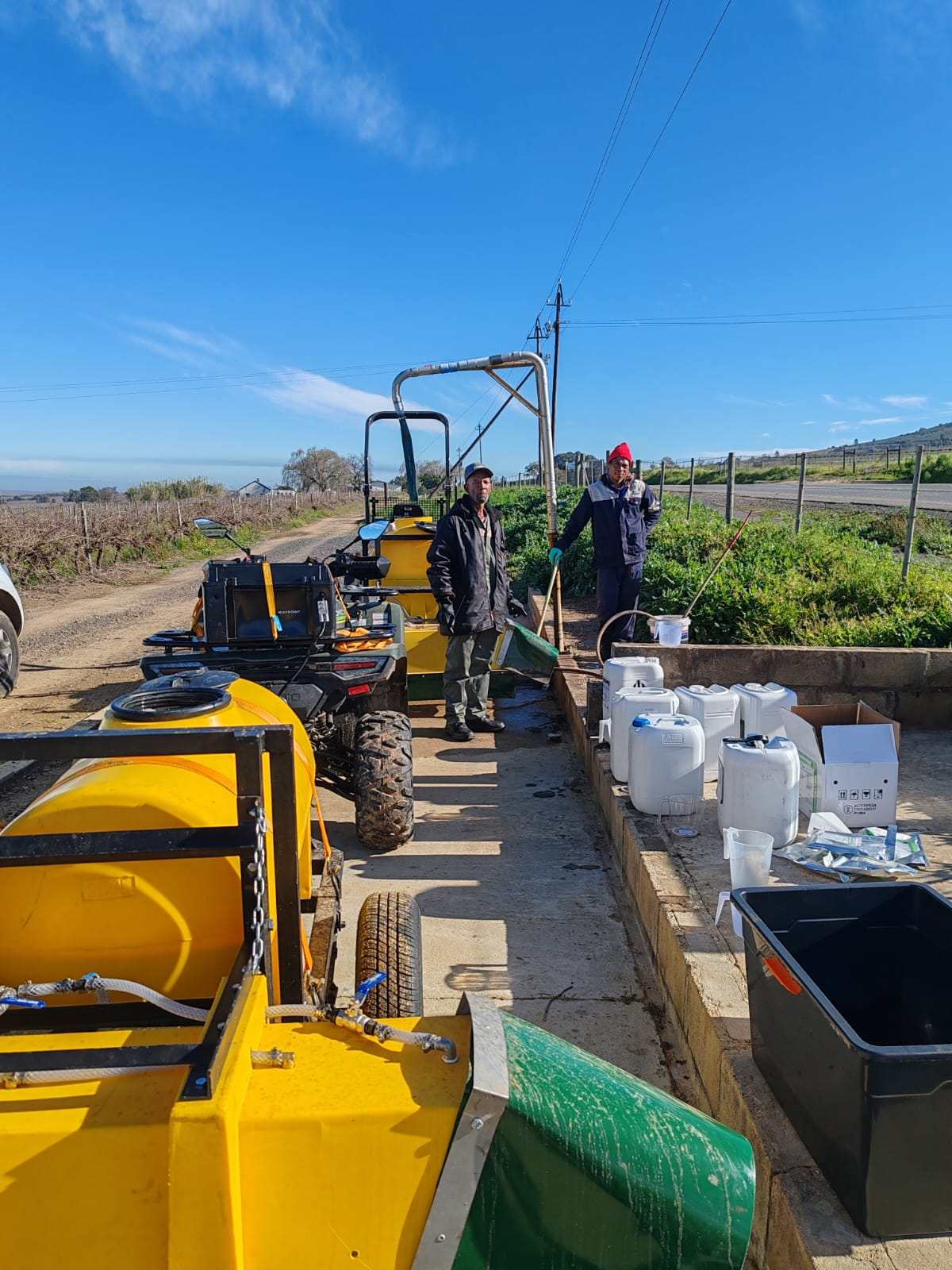Two crew members preparing herbicide application beside the yellow spray tank in a vineyard, with chemical containers ready on a sunny day.