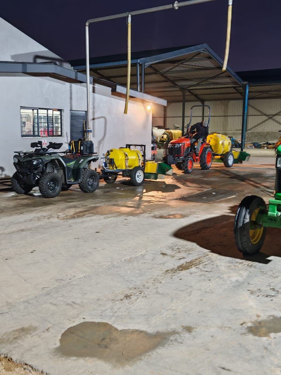 Quad bike, small Kubota tractor, and trailed equipment staged in the depot shed at dusk.