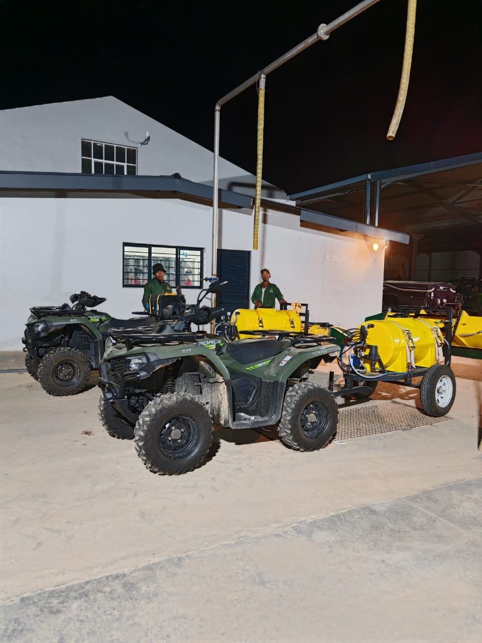 Two ATV quad bikes hitched to yellow-tank pesticide sprayers under cover at the equipment depot, ready for a programme run.