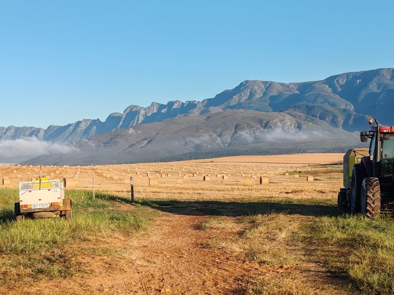 Tractor and trailer at the edge of a freshly-baled field with the Langeberg mountains catching morning light.