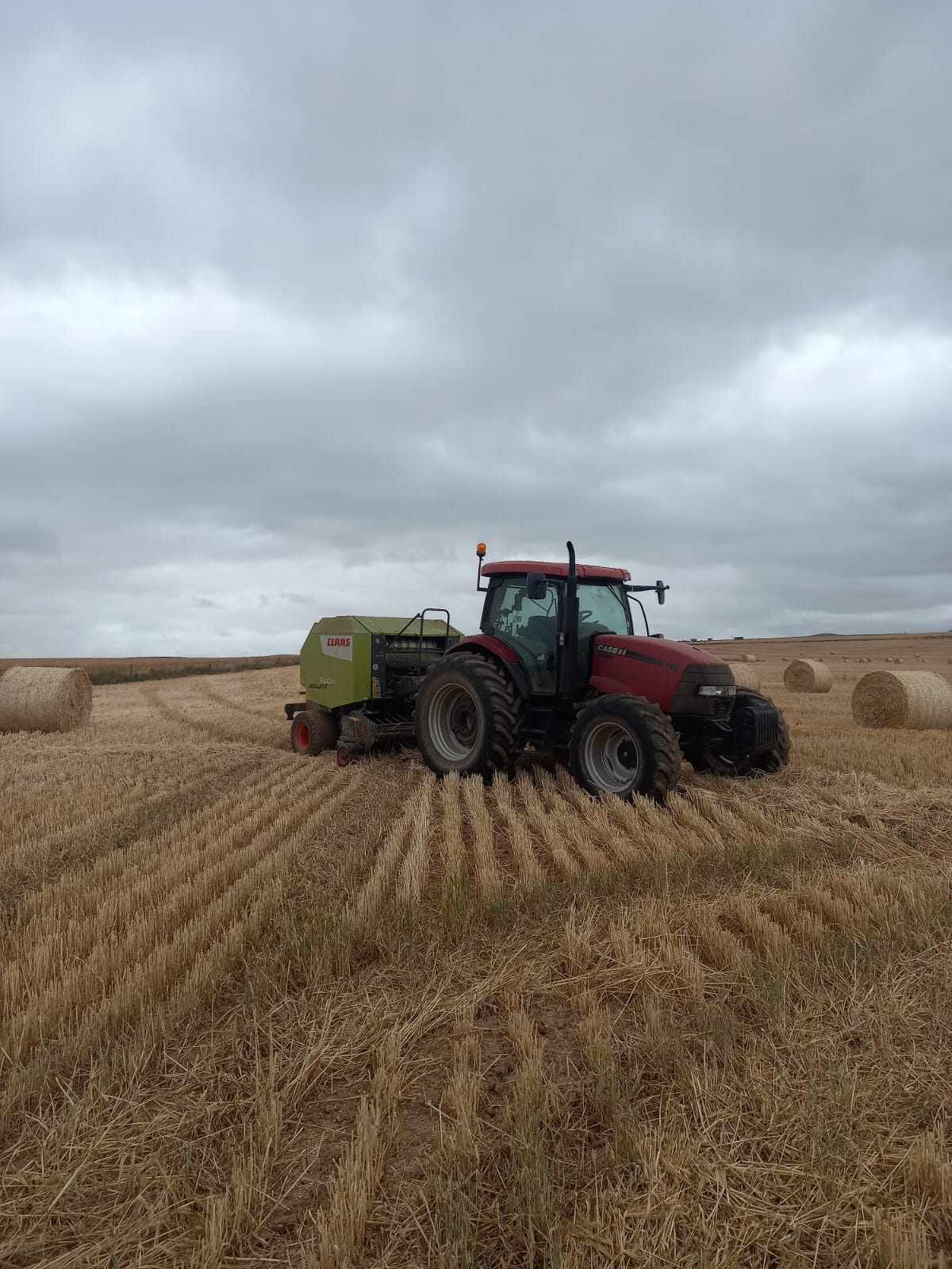 Red CASE tractor towing a green CLAAS round baler through a wheat stubble field on a grey day, with finished round bales in the background.