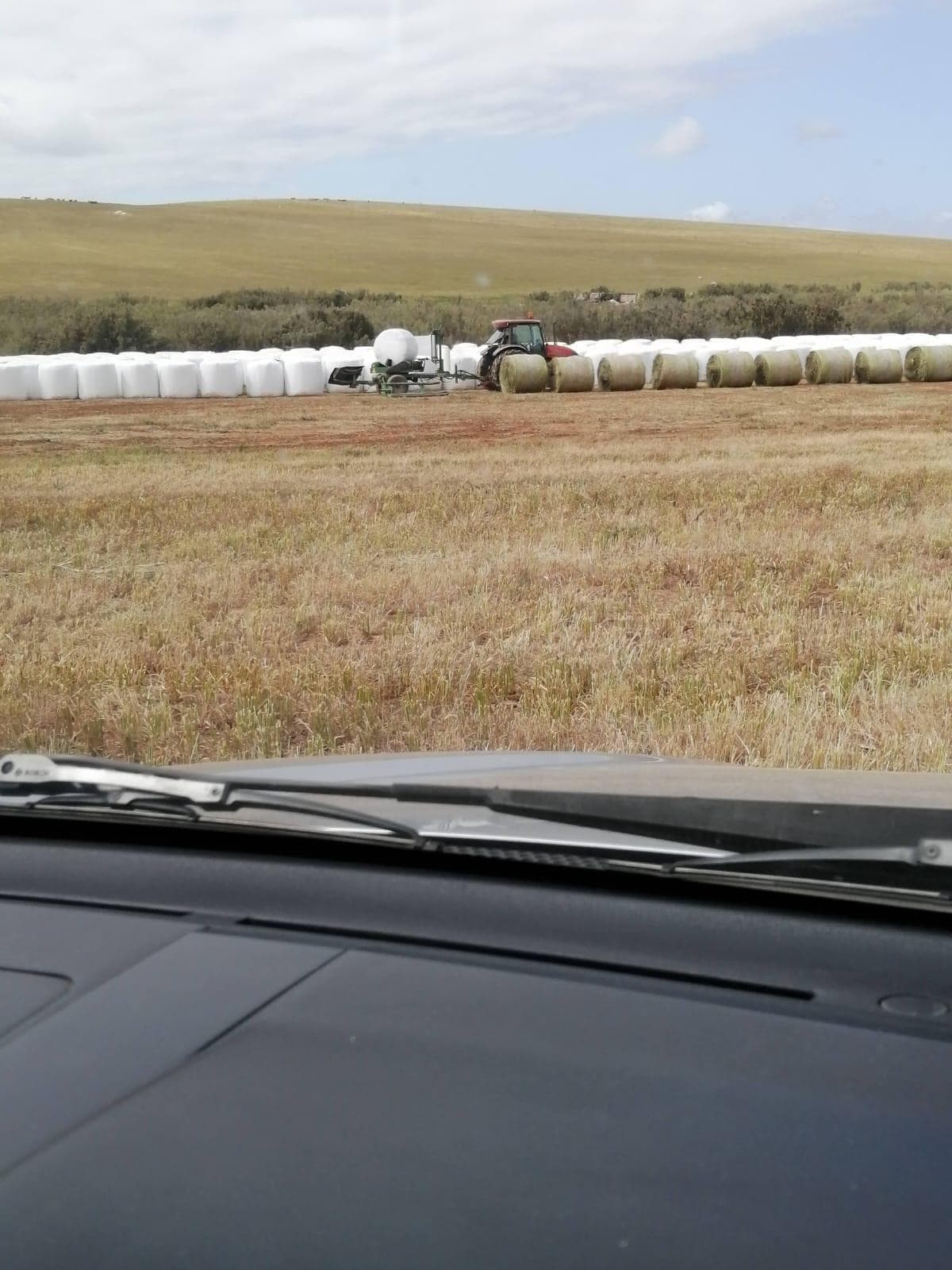 View from a vehicle cab of a tractor wrapping silage bales in white plastic at the edge of a baled field.