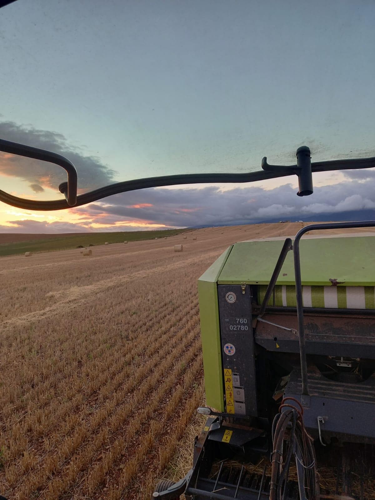 View across a stubble field at sunset from the cab of a tractor with a CLAAS round baler attached.